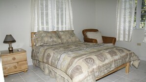 A bedroom with a bed, a nightstand, a lamp, and a wicker chair. - Friars Hill Cottage (St. John's)