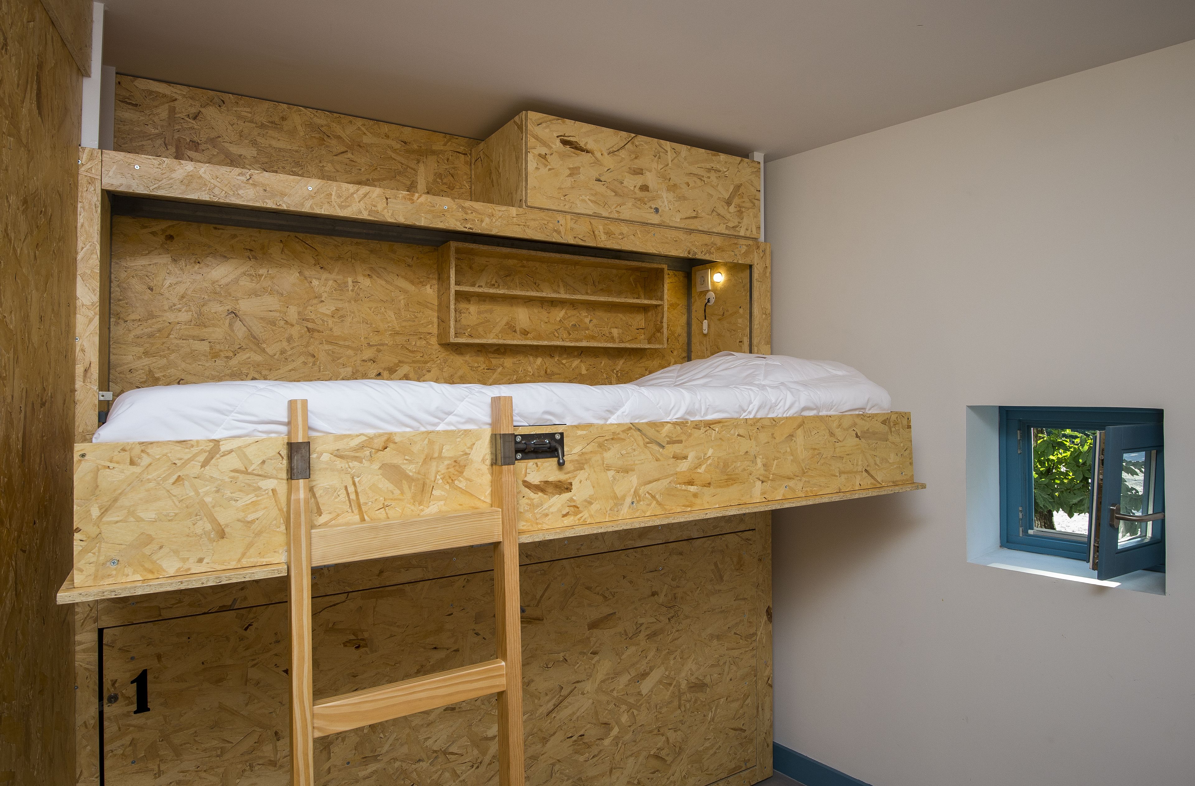 A bunk bed with a ladder, a small shelf, and a window with a view of greenery.