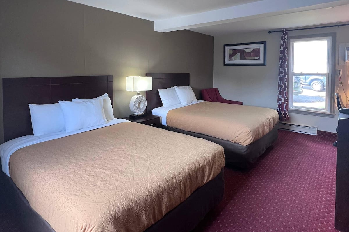 A hotel room with two beds, a nightstand with a lamp, a window with a view of a car, and a door with a curtain.