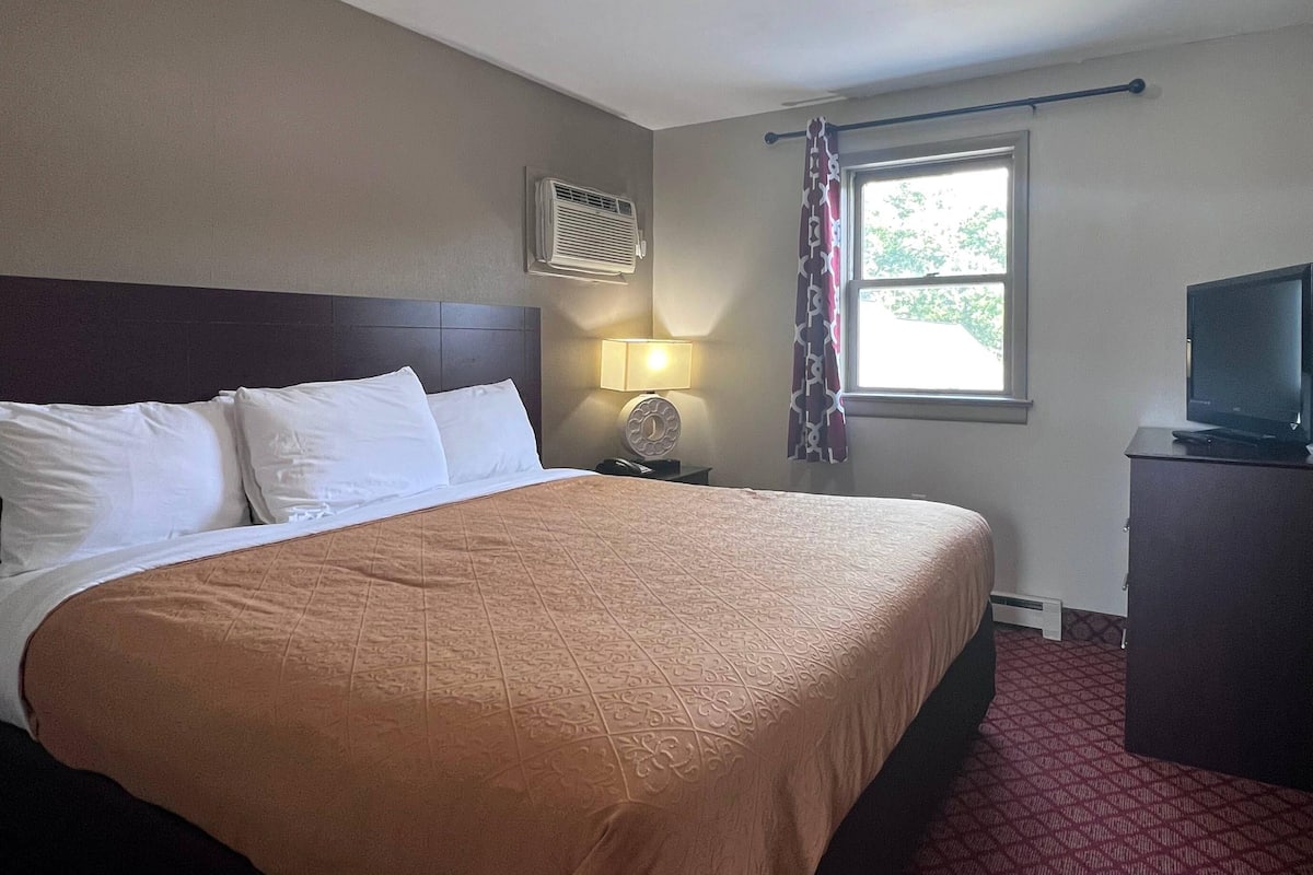 A hotel room with a large bed, a bedside lamp, an air conditioning unit, a television on a cabinet, and a window with curtains.