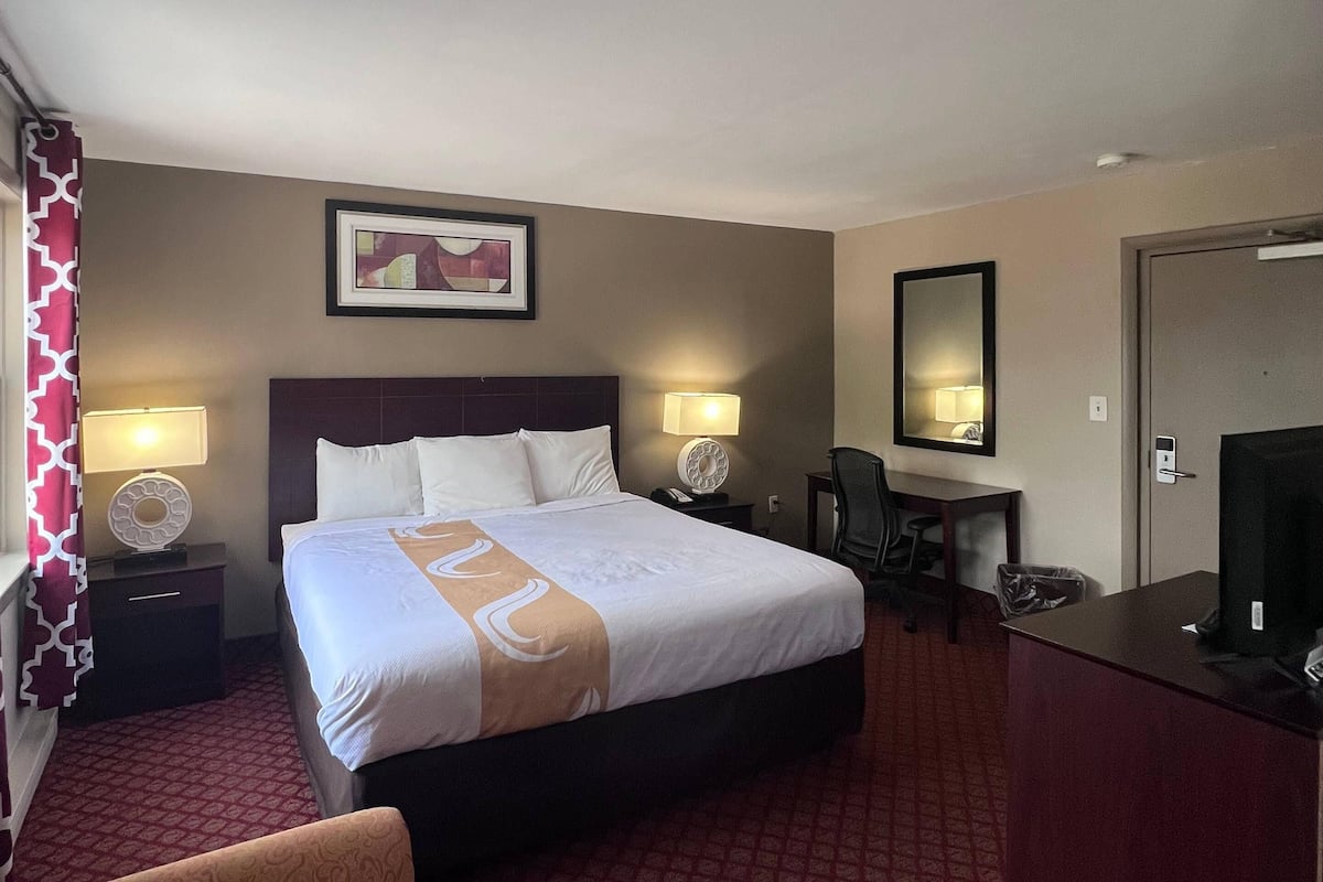 A hotel room with a large bed, two bedside tables with lamps, a desk with a chair, a mirror, and a red patterned carpet.