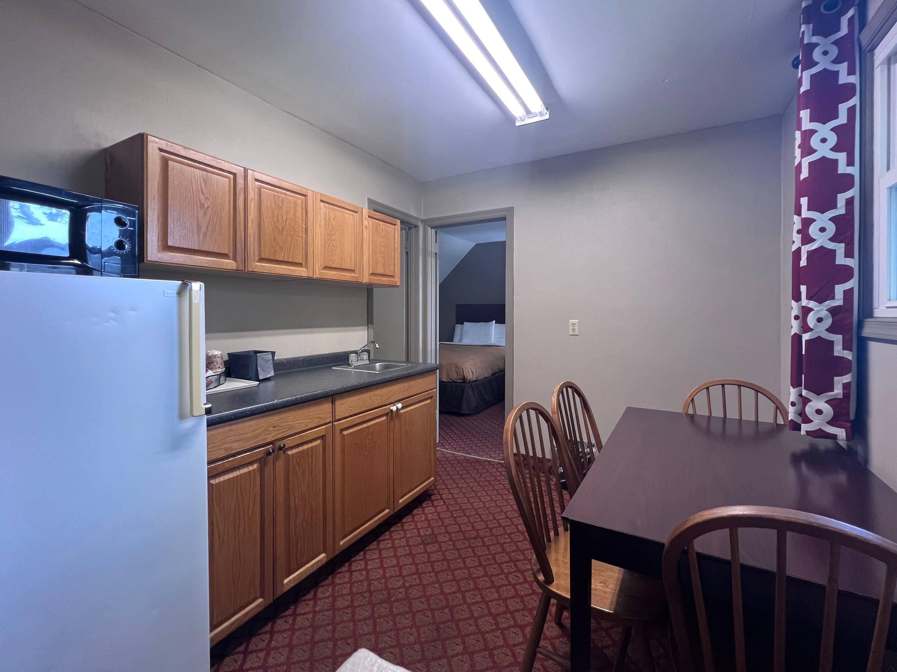 Una cocina compacta con armarios de madera, un refrigerador y un microondas. Hay una zona de comedor con una mesa y sillas, y una alfombra estampada.