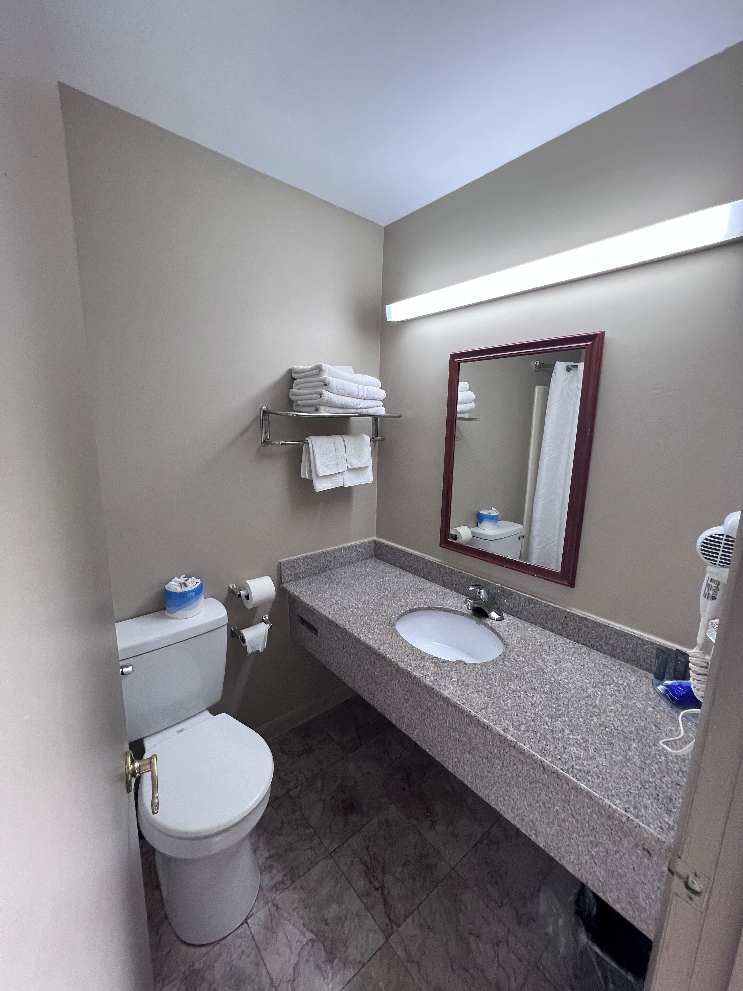 Casa de banho com sanita, lavatório e espelho. Há toalhas numa prateleira e um secador de cabelo sobre a bancada.
