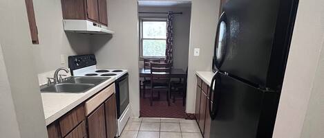 A compact kitchen with wooden cabinets, a black refrigerator, a stove, and a window with curtains.