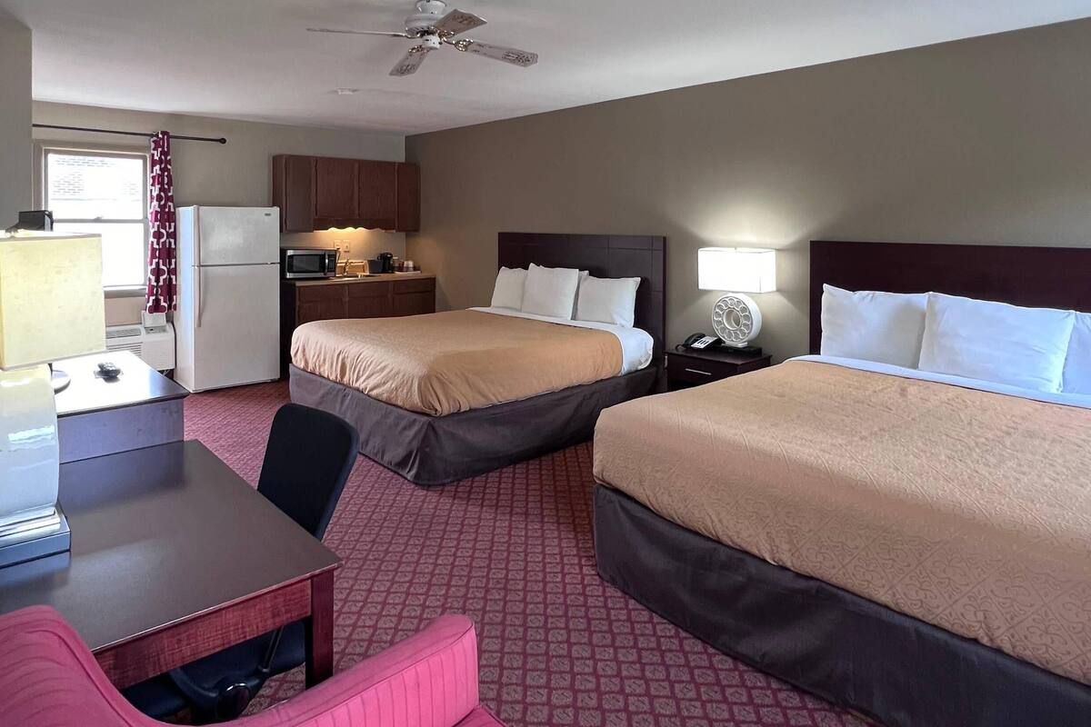 A hotel room with two beds, a desk, a chair, a kitchenette, and a ceiling fan.