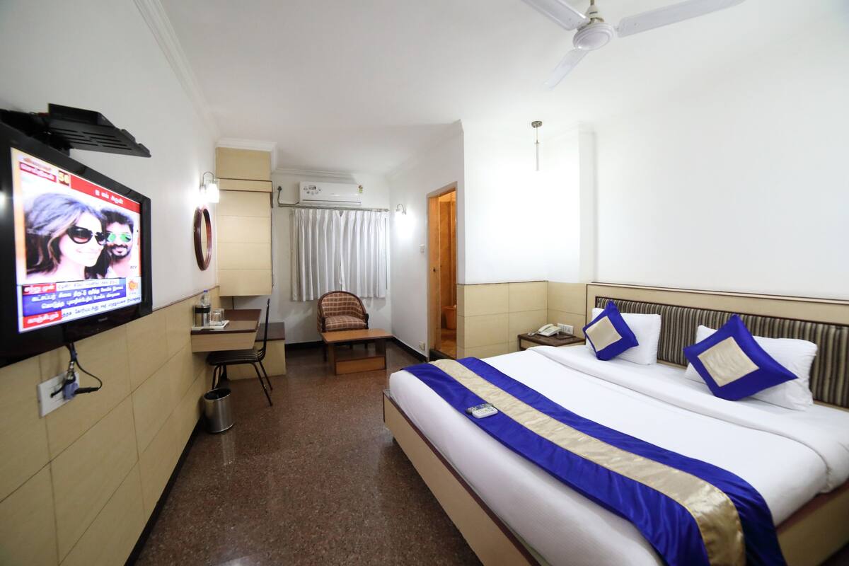 A hotel room with a bed, a desk, a chair, a television, and an air conditioning unit.