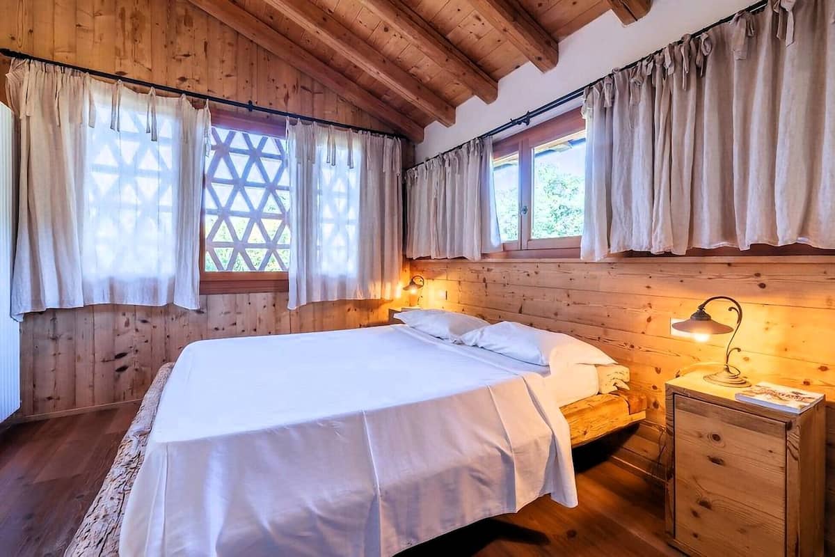 A wooden cabin bedroom with a bed, two windows with curtains, a bedside lamp, and a wooden nightstand.
