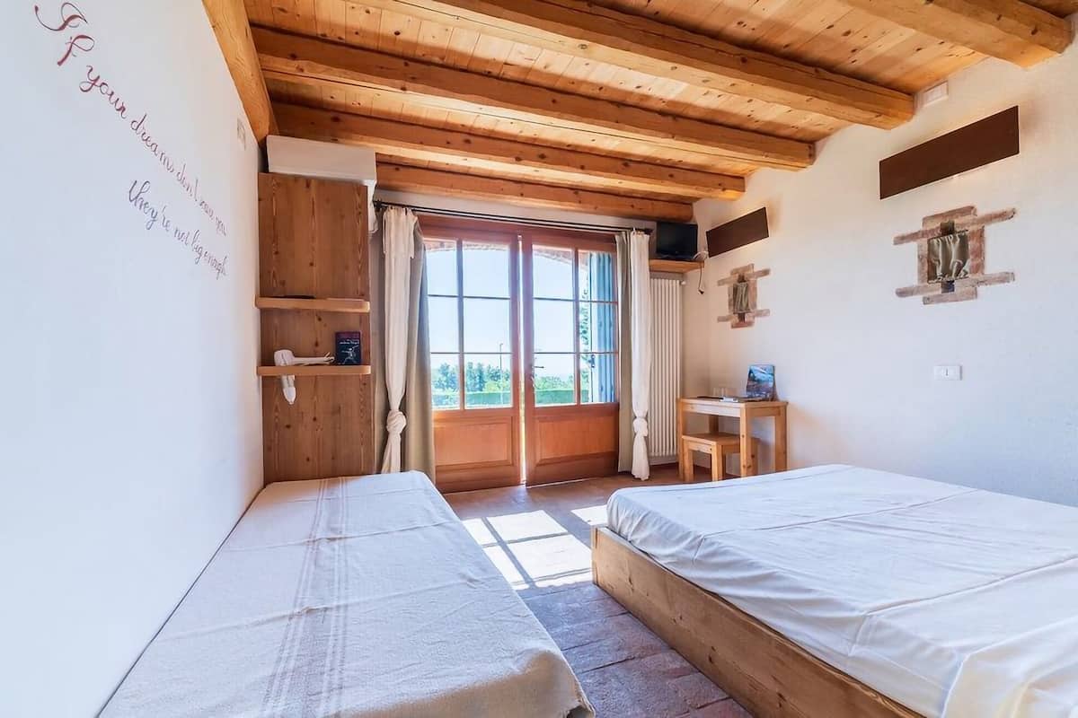 A bedroom with two beds, wooden ceiling beams, a window with curtains, a small table, and a shelf with items.