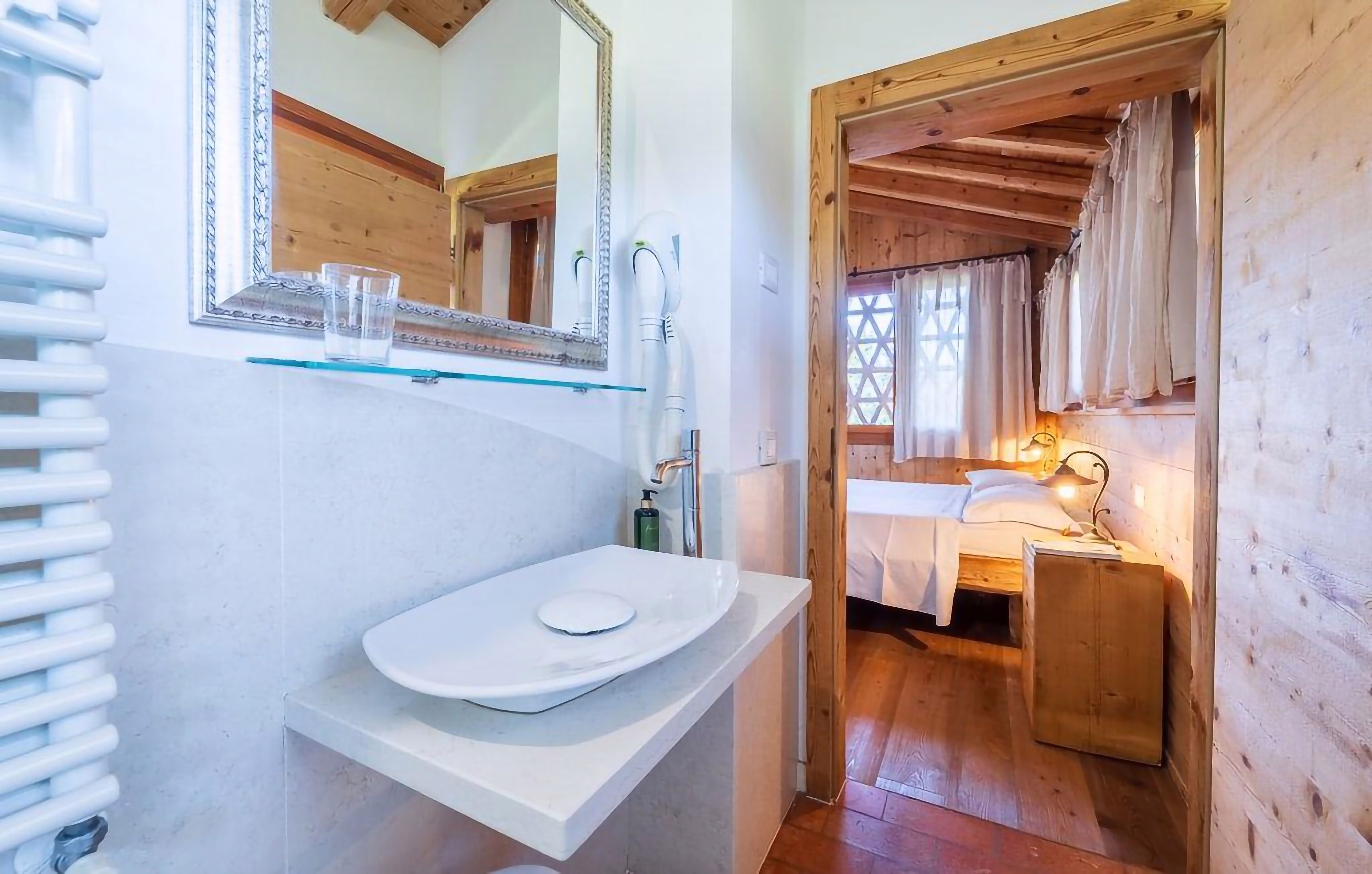 A bathroom with a white sink, mirror, and radiator. There is a wooden door leading to a bedroom with a bed and a nightstand.