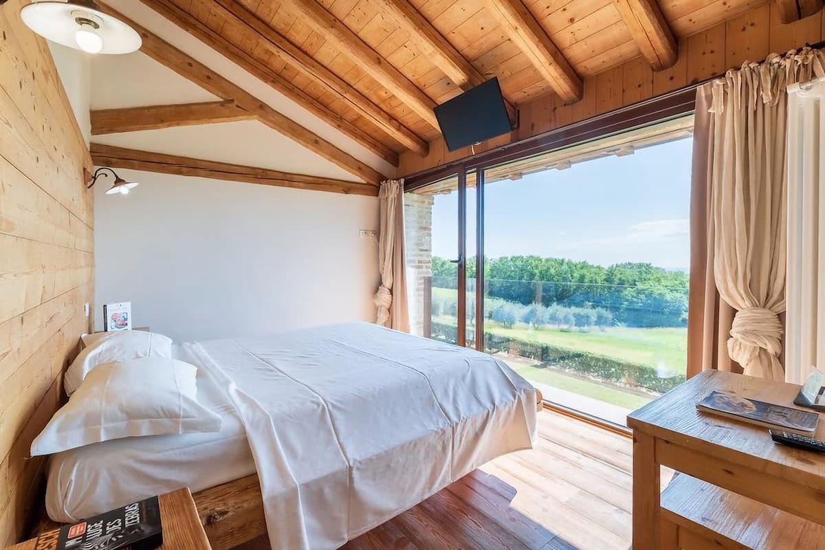 A bedroom with a bed, a table, and a large window with a view of greenery.