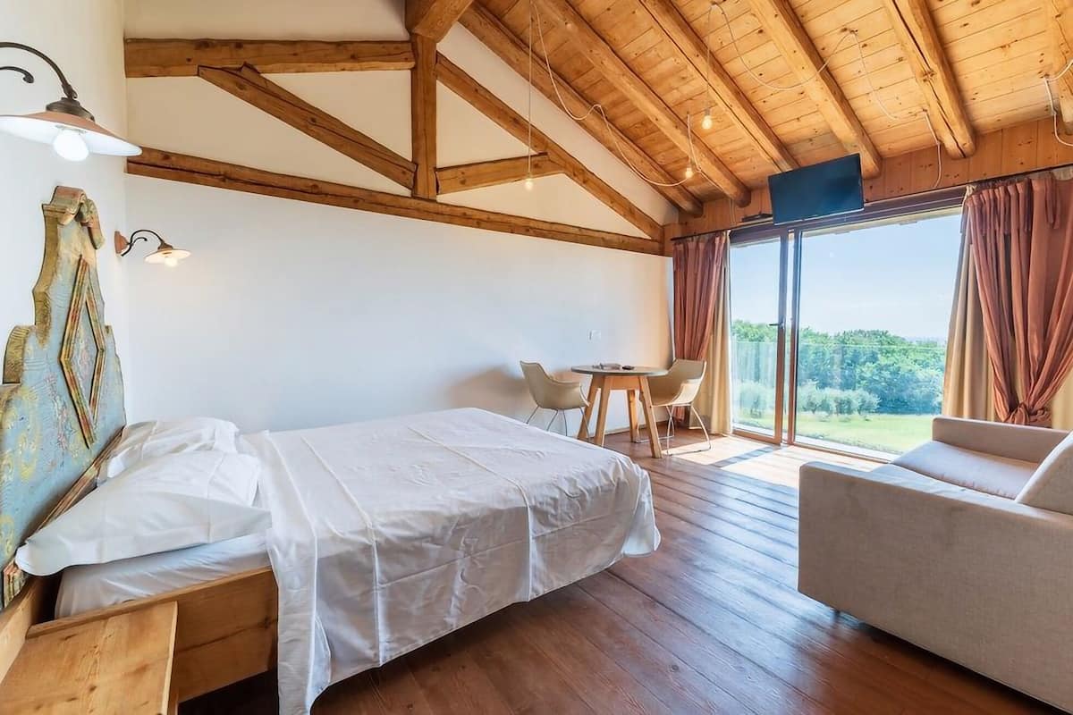 A bedroom with a large bed, a small table with chairs, and a view of the outdoors through a sliding door.