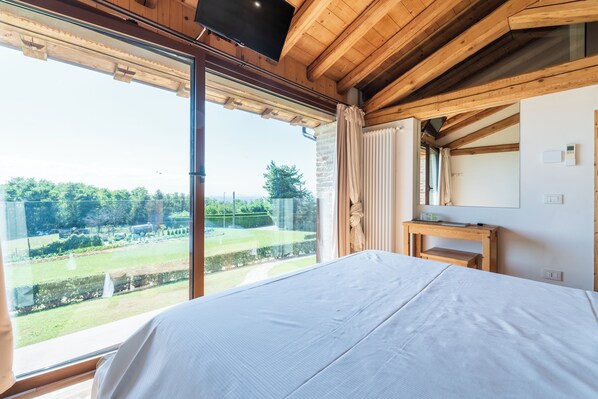 A bedroom with a large window, wooden ceiling beams, a bed, a small wooden table, and a TV. - Agriturismo Dolce Colle (Volpago del Montello)