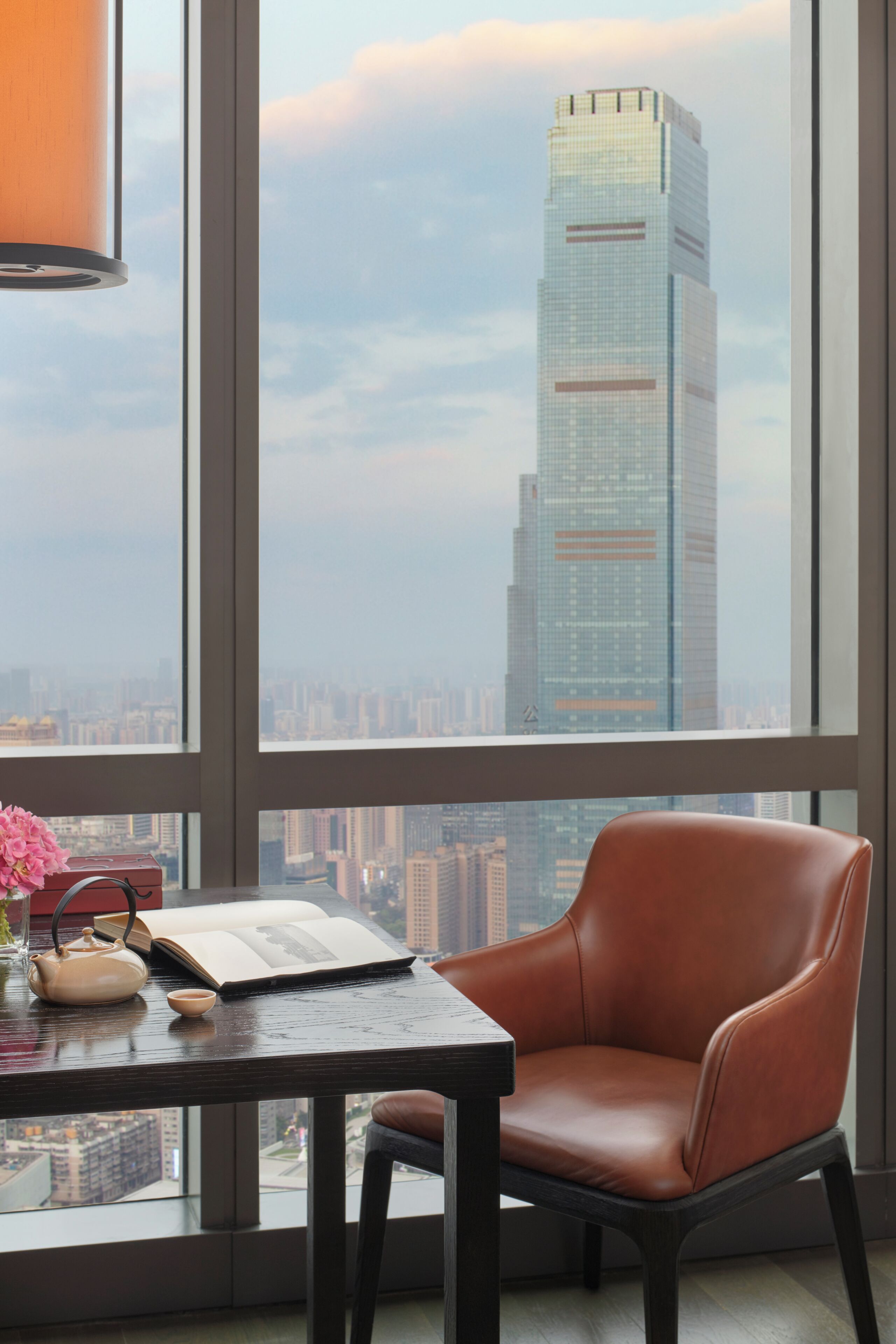 A modern room with a large window, a leather chair, a table with a book and a teapot, and a view of a tall building.