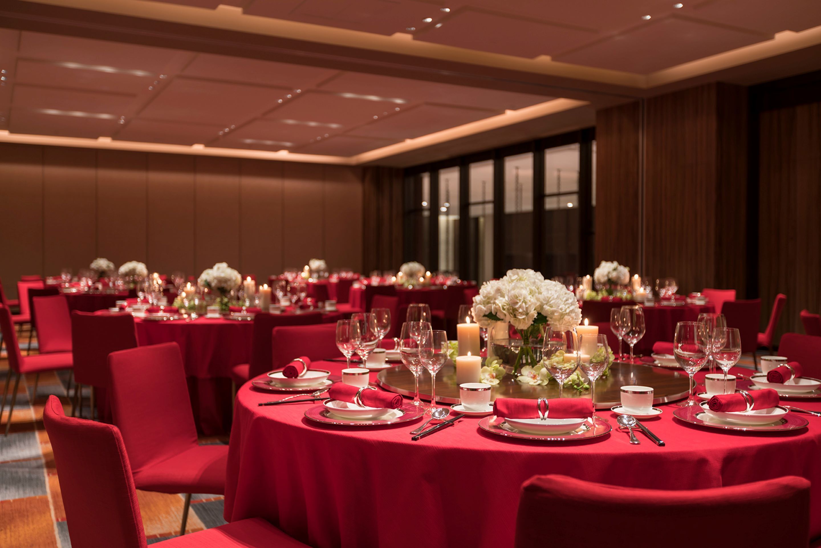 Un salón de banquetes con mesas redondas dispuestas para el servicio de comidas, cada una con manteles rojos, platos blancos y servilletas rojas.