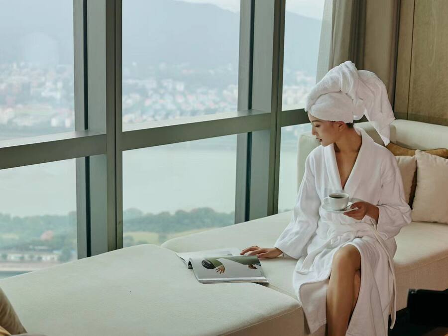 A person in a white bathrobe sitting on a sofa with a book and a cup, overlooking a cityscape through large windows.