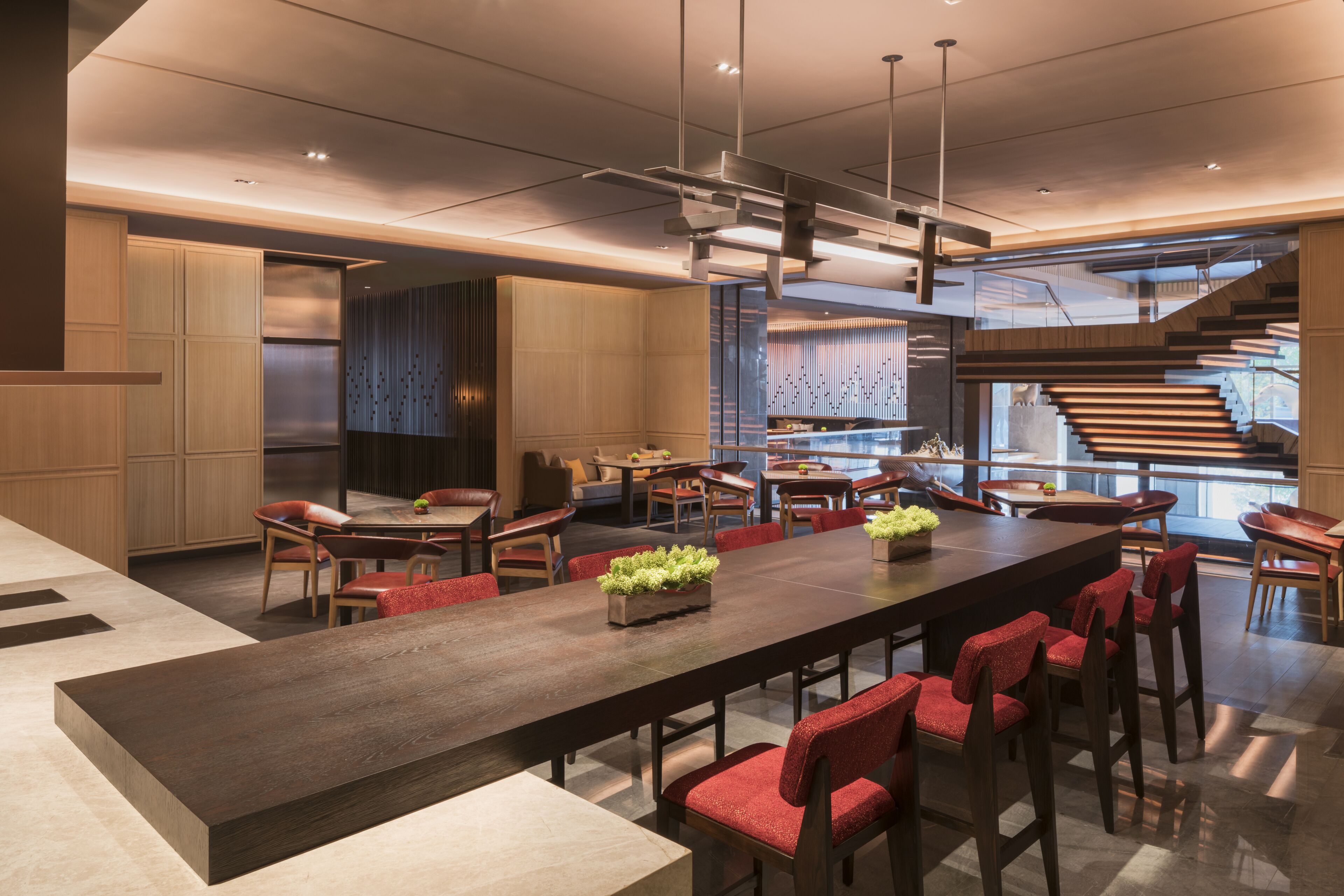 A modern dining area with a long table, red upholstered chairs, and a central plant.