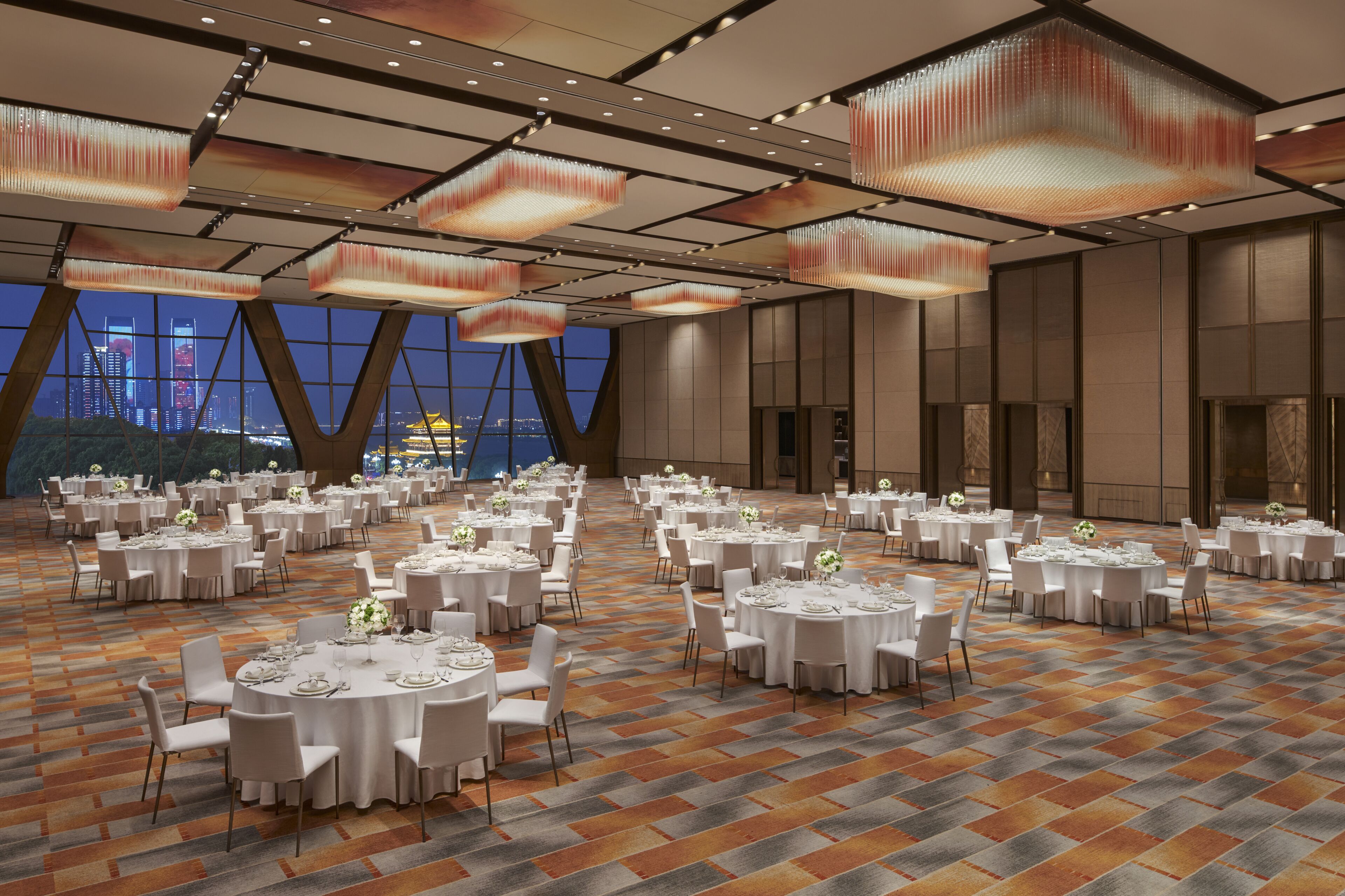 A large banquet hall with round tables set for dining, featuring a patterned carpet and modern ceiling lights.