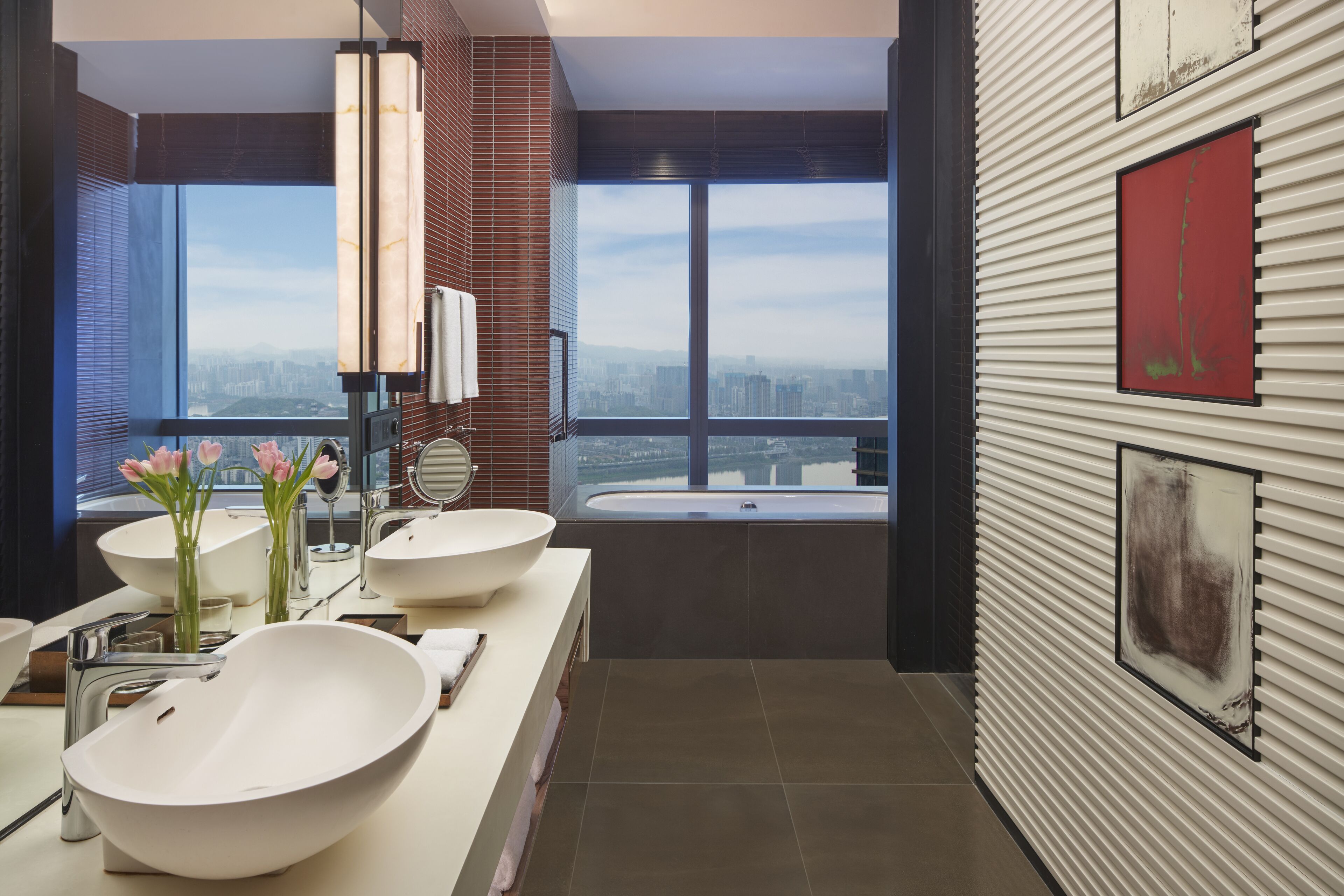 Un baño moderno con dos lavamanos grandes blancos, un espejo amplio y una bañera. Amplias ventanas ofrecen vistas a la ciudad.