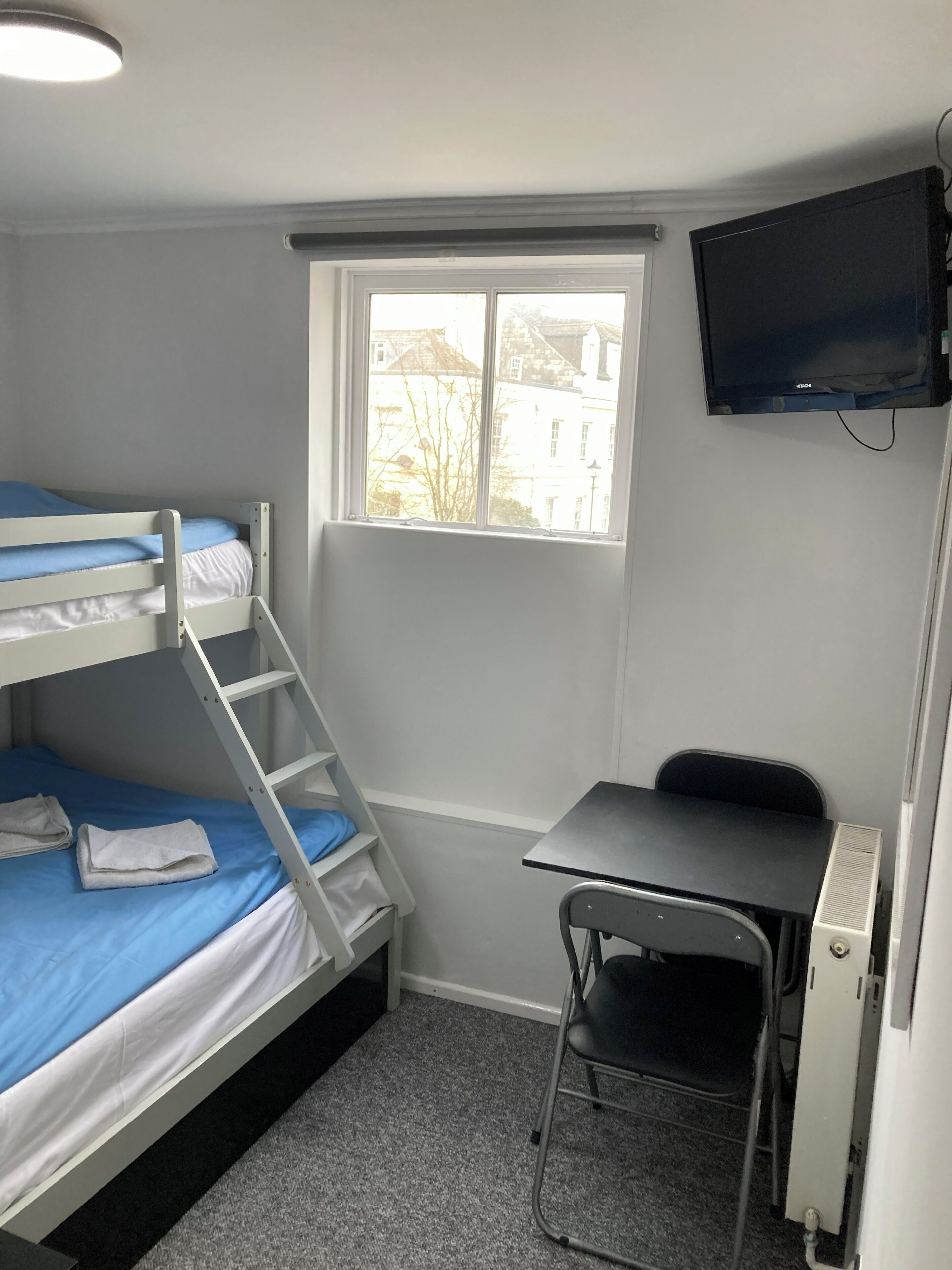 A dormitory room with bunk beds, a desk, a chair, a television, and a window with a view of buildings.
