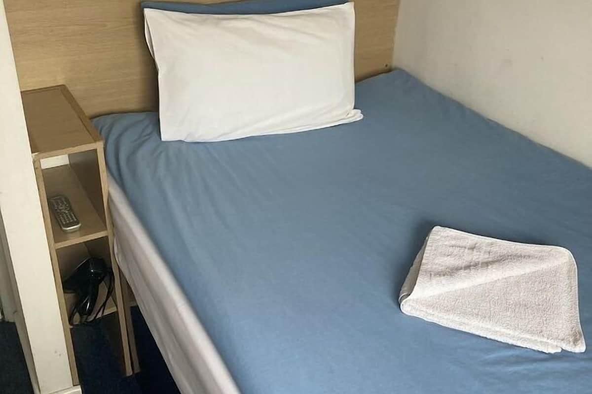 A single bed with a blue mattress and white pillows, a wooden headboard, a bedside table with a lamp, and a folded towel on the bed.