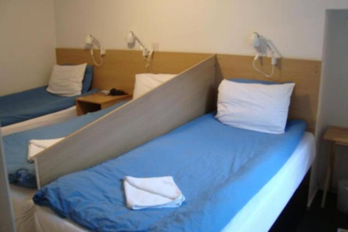 A hotel room with two single beds, each with a blue mattress and white pillows. There are bedside tables with lamps and a small wooden nightstand.