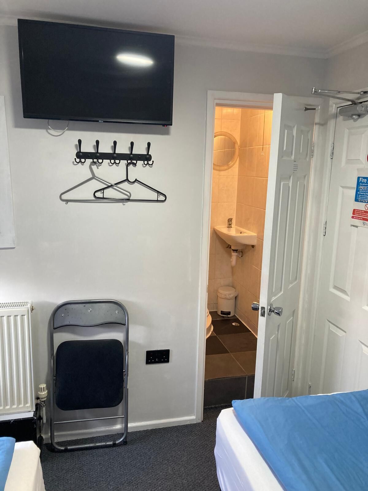 Habitación de hotel con cama, televisor montado en la pared, perchero, radiador y baño visible a través de una puerta abierta.