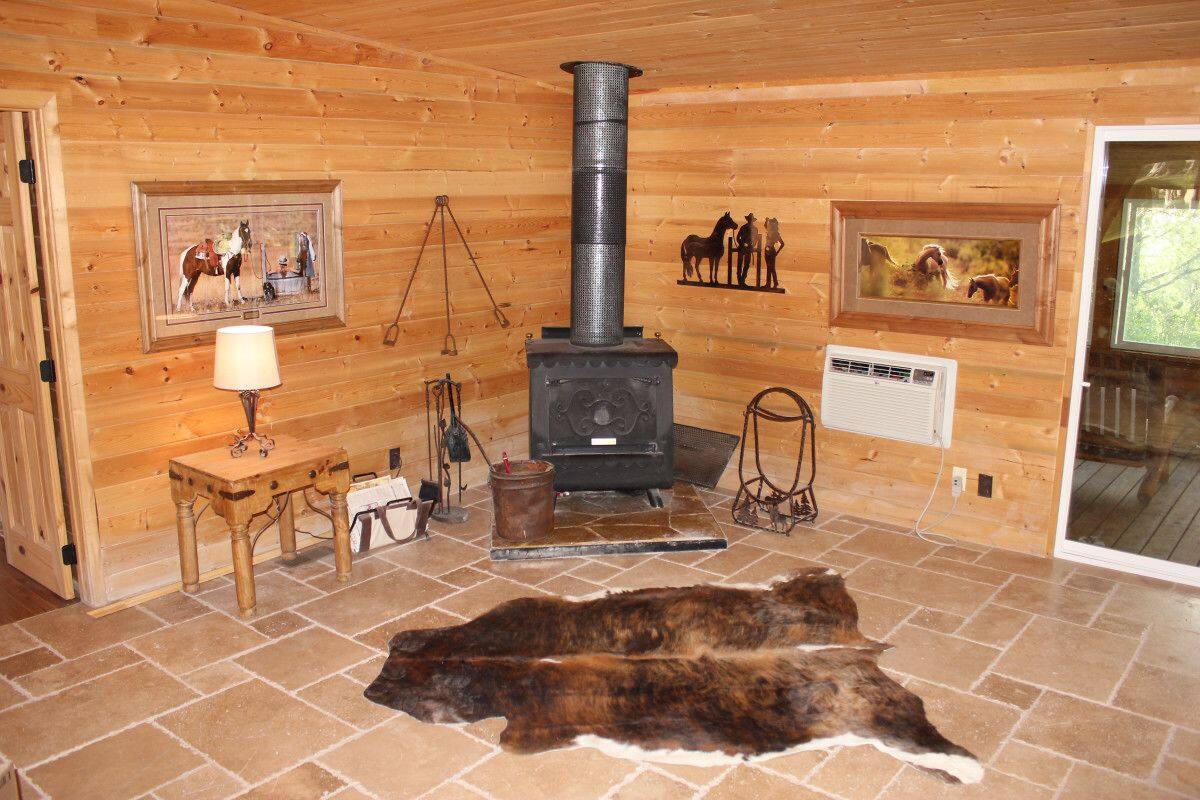 A rustic cabin interior with a wood-burning stove, framed equestrian paintings, a leather hide on the floor, and a small table with a lamp.