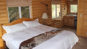 A log cabin bedroom with a wooden bed, a nightstand, a lamp, a small table, and a window with a view of trees.