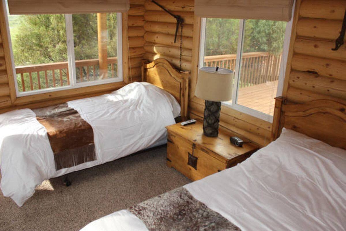A cabin with two single beds, a wooden nightstand, a lamp, and a view of a deck and trees.
