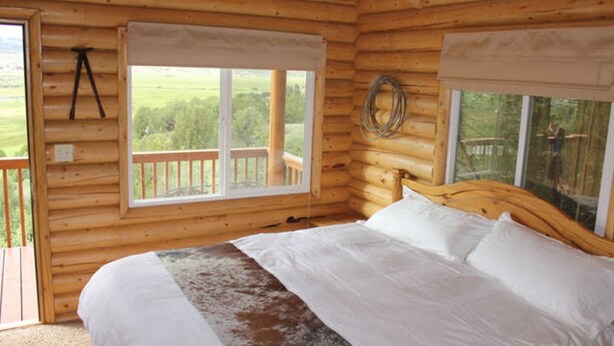 A wooden cabin bedroom with a large bed, two windows with blinds, a wooden headboard, and a balcony with a railing.