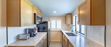 A modern kitchen with wooden cabinets, a stainless steel refrigerator, a built-in oven, and a window with blinds.