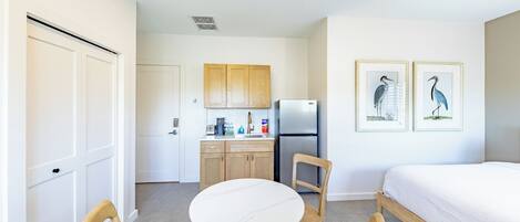 A compact room with a bed, a dining table with chairs, a kitchenette, and two framed pictures on the wall.