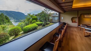 A room with a long counter, wooden chairs, and a view of a lush garden and mountains. - Washintei Hogetsu (Hakone)