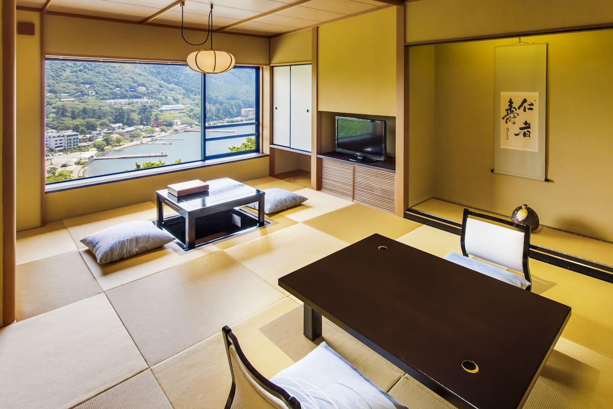 A modern room with tatami flooring, a low table, a sofa, a TV, and a view of a river and hills.
