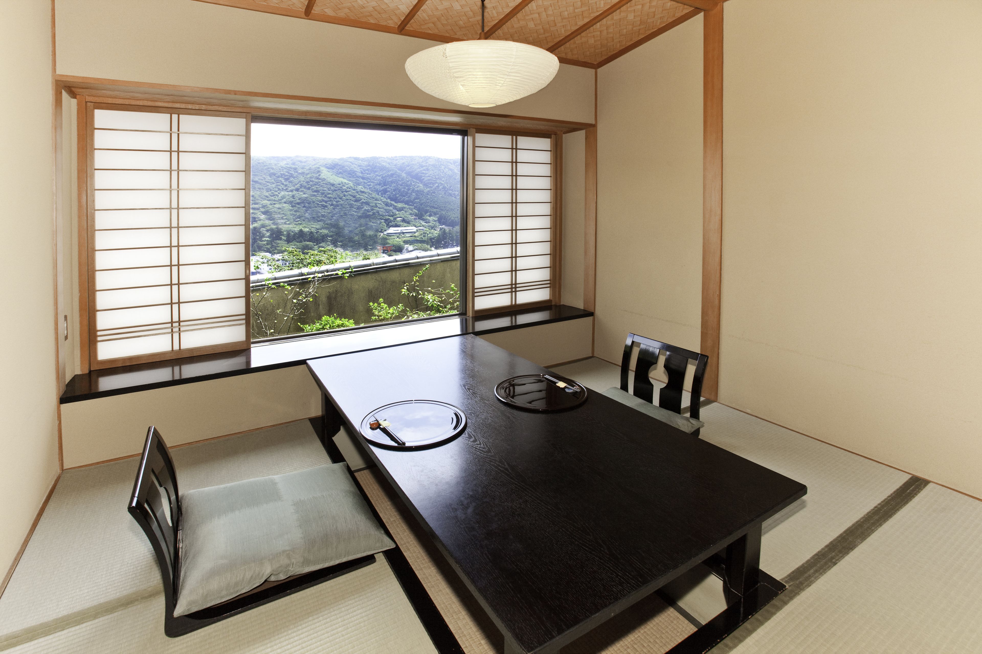 A traditional Japanese room with tatami flooring, a black table with two tatami mats, a low wooden bench, and a view of a mountainous landscape through shoji screens.