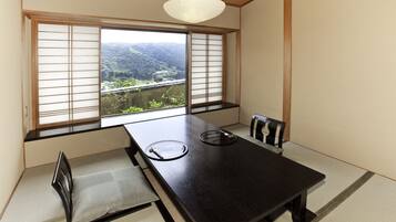 A traditional Japanese room with tatami flooring, a black table with two tatami mats, a low wooden bench, and a view of a mountainous landscape through shoji screens.