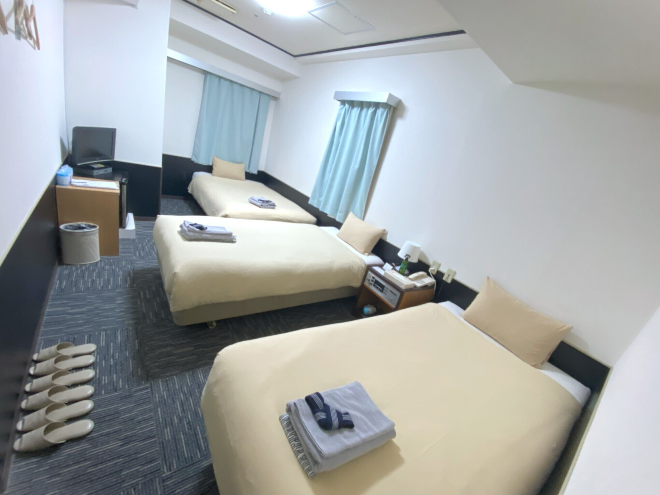 A hotel room with two beds, a desk, a chair, a trash bin, and a window with curtains.
