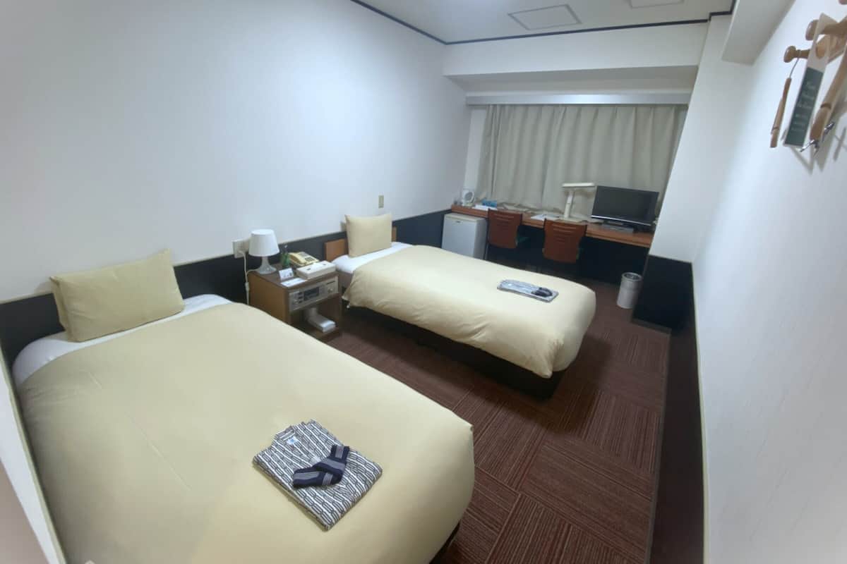 A hotel room with two beds, a desk with a computer, a small refrigerator, and a window with curtains.
