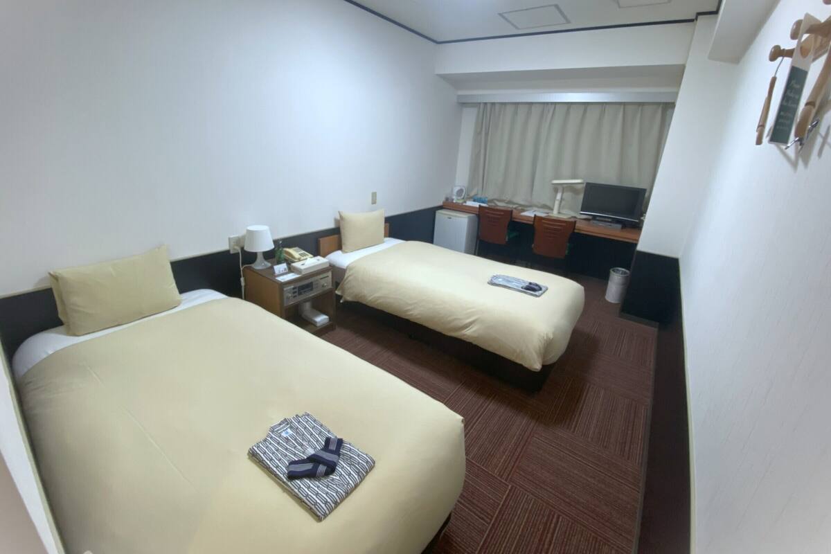 Habitación de hotel con dos camas, un escritorio con computadora, un refrigerador pequeño y una ventana con cortinas.