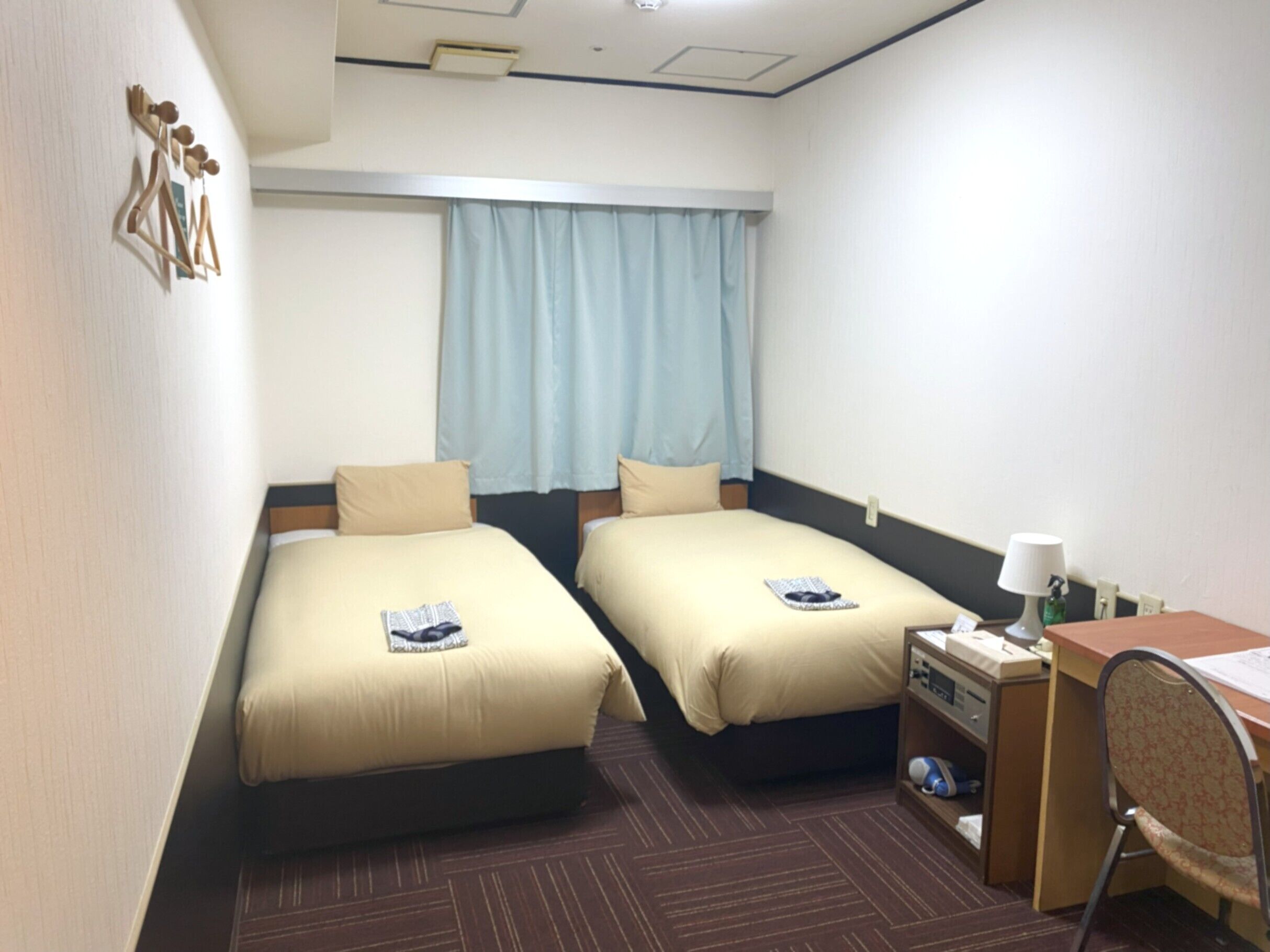A hotel room with two beds, a desk, a chair, a lamp, and a window with curtains.