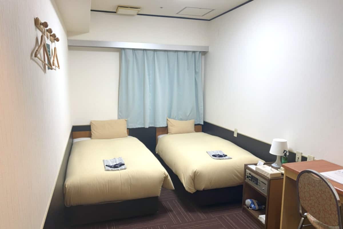 Habitación de hotel con dos camas, un escritorio, una silla, una lámpara y una ventana con cortinas.