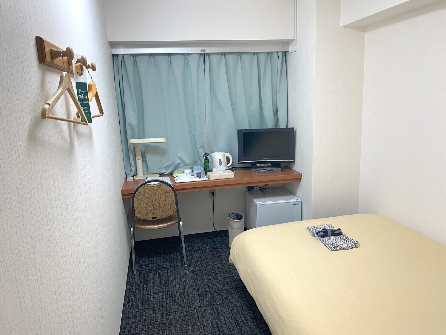 A hotel room with a bed, a desk with a television, a chair, a small refrigerator, and a window with curtains.