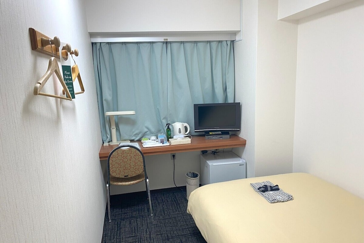 A hotel room with a bed, a desk with a television, a chair, a small refrigerator, and a window with curtains.