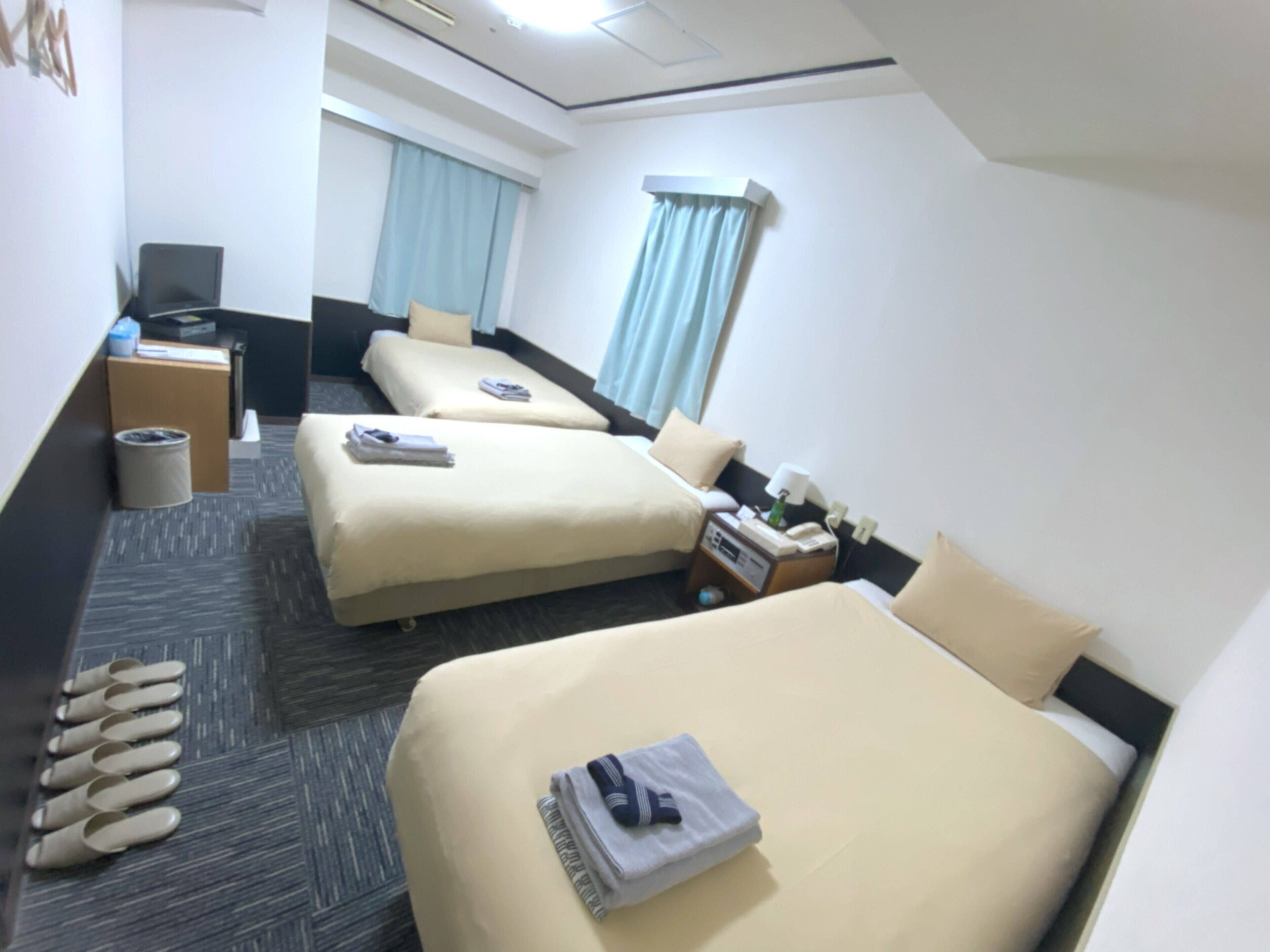 A hotel room with two beds, a desk, a chair, a trash bin, and a window with curtains.
