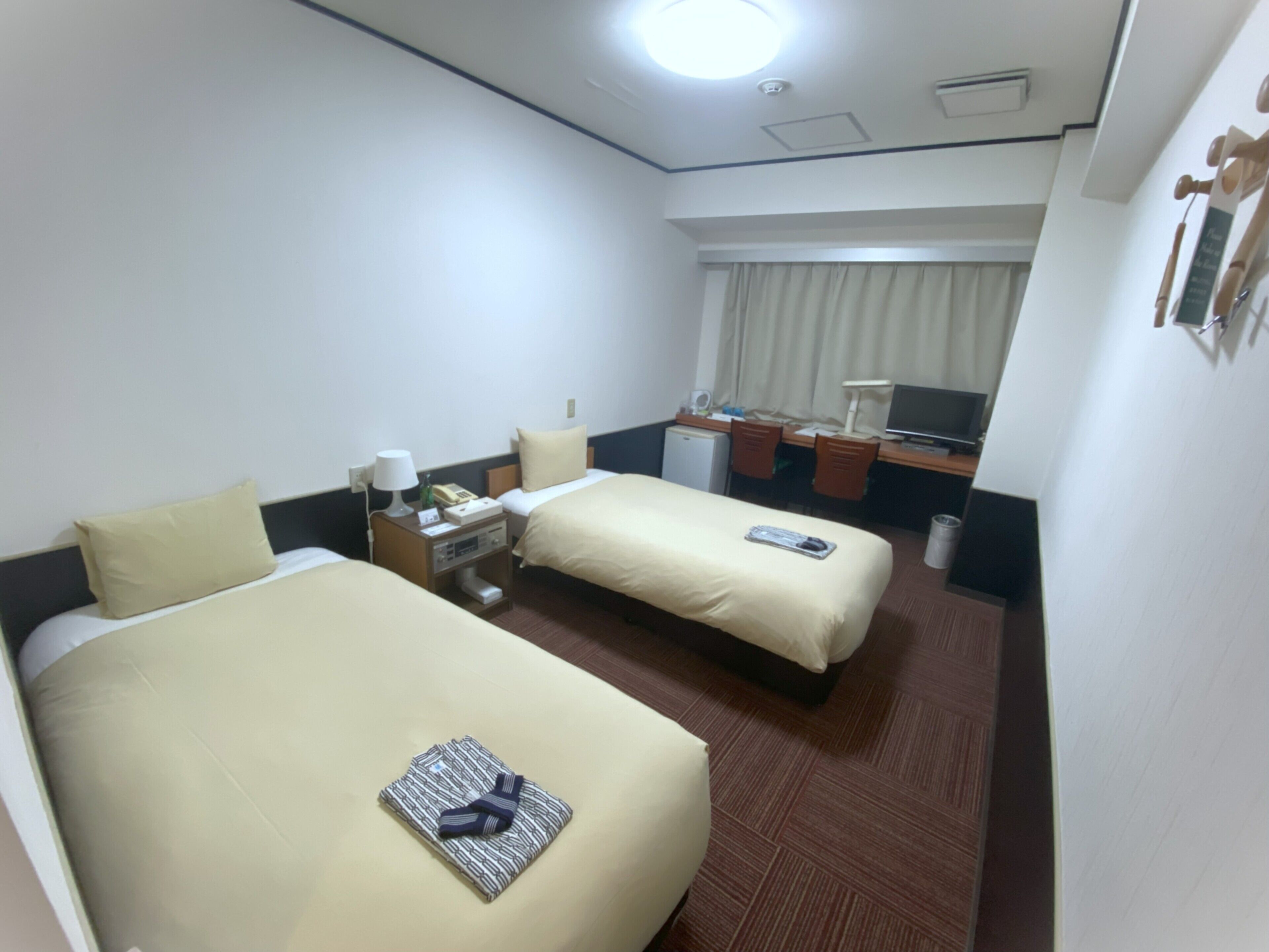 A hotel room with two beds, a desk with a chair, a small refrigerator, and a window with curtains.