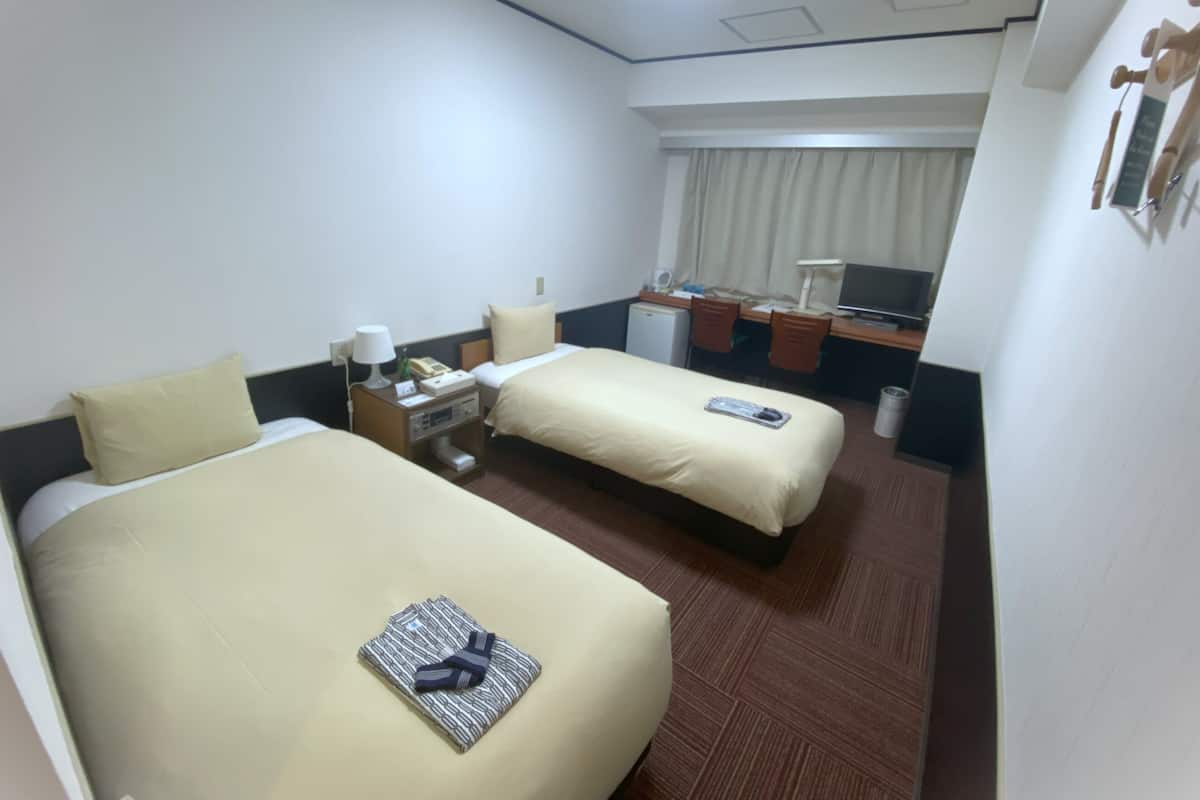 A hotel room with two beds, a desk with a chair, a small refrigerator, and a window with curtains.