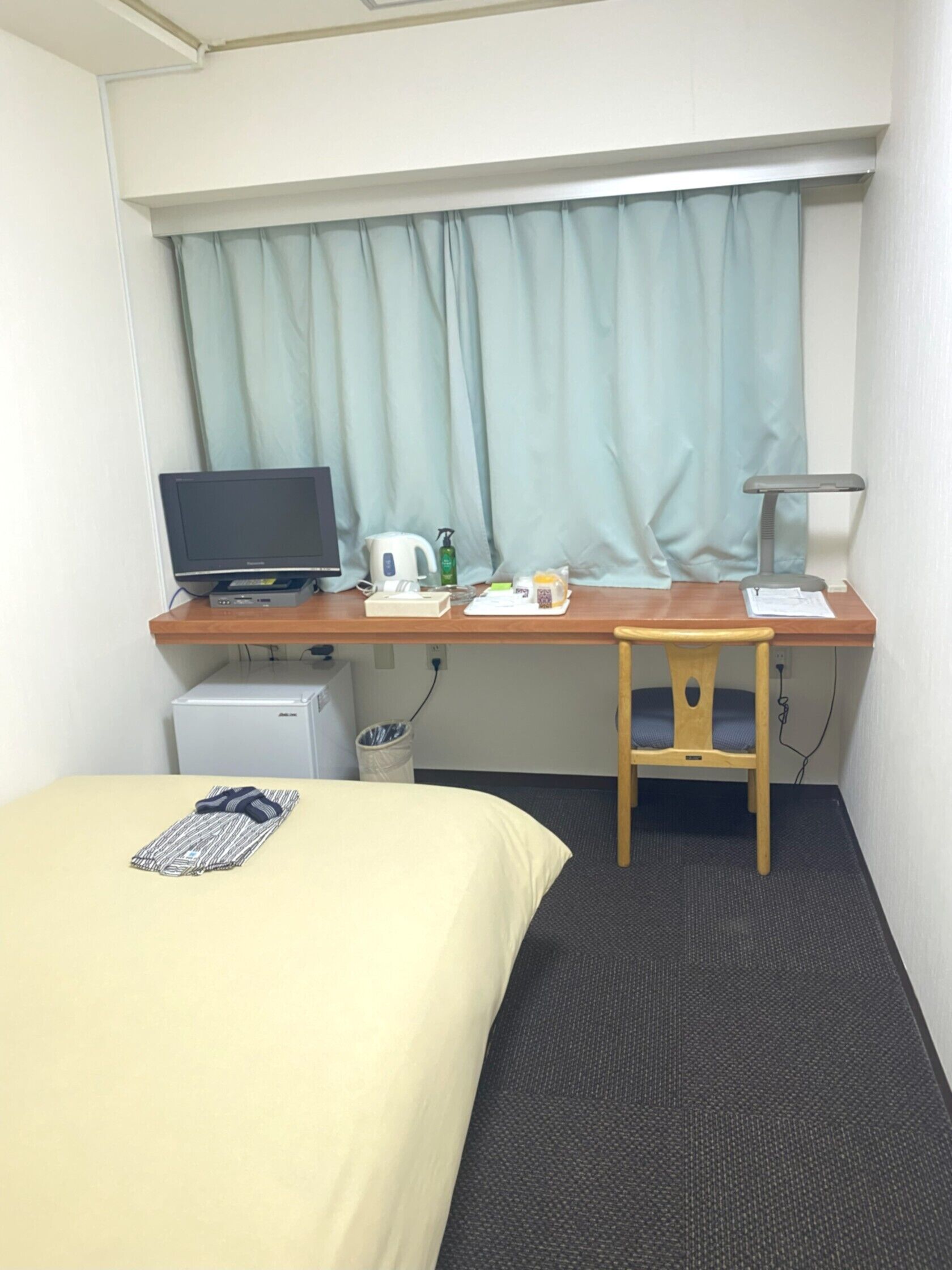 A hotel room with a bed, a desk with a computer, a chair, a small fridge, and a window with curtains.