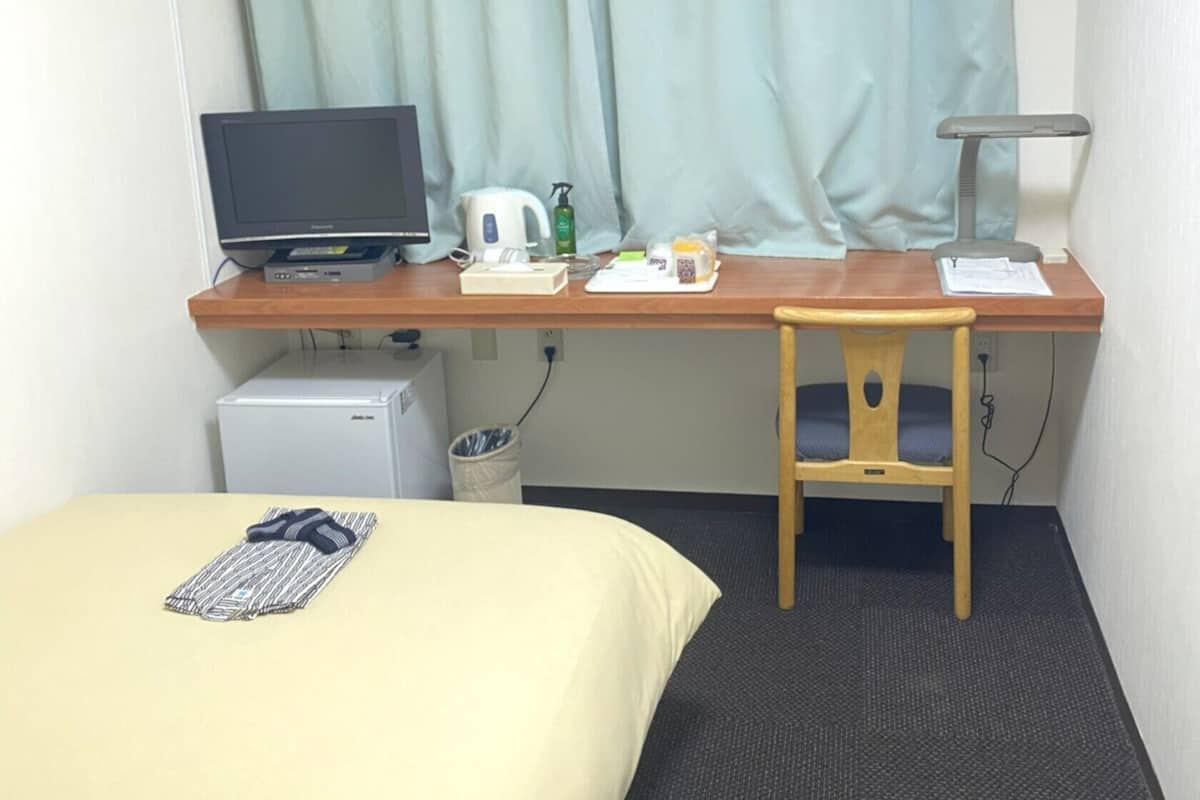 A hotel room with a bed, a desk with a computer, a chair, a small fridge, and a window with curtains.