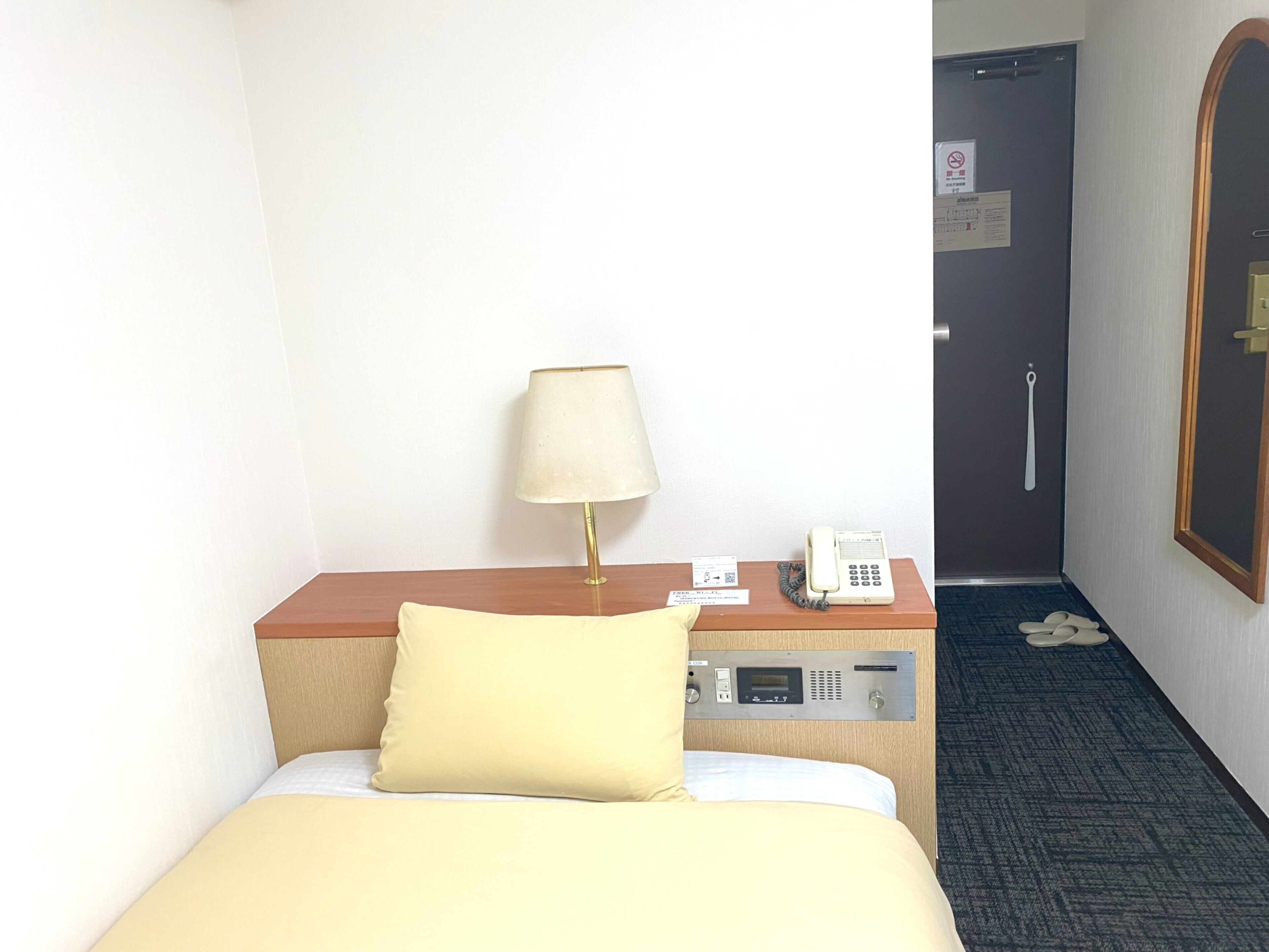 A hotel room with a bed, a bedside table with a lamp, a phone, and a door with a handle.