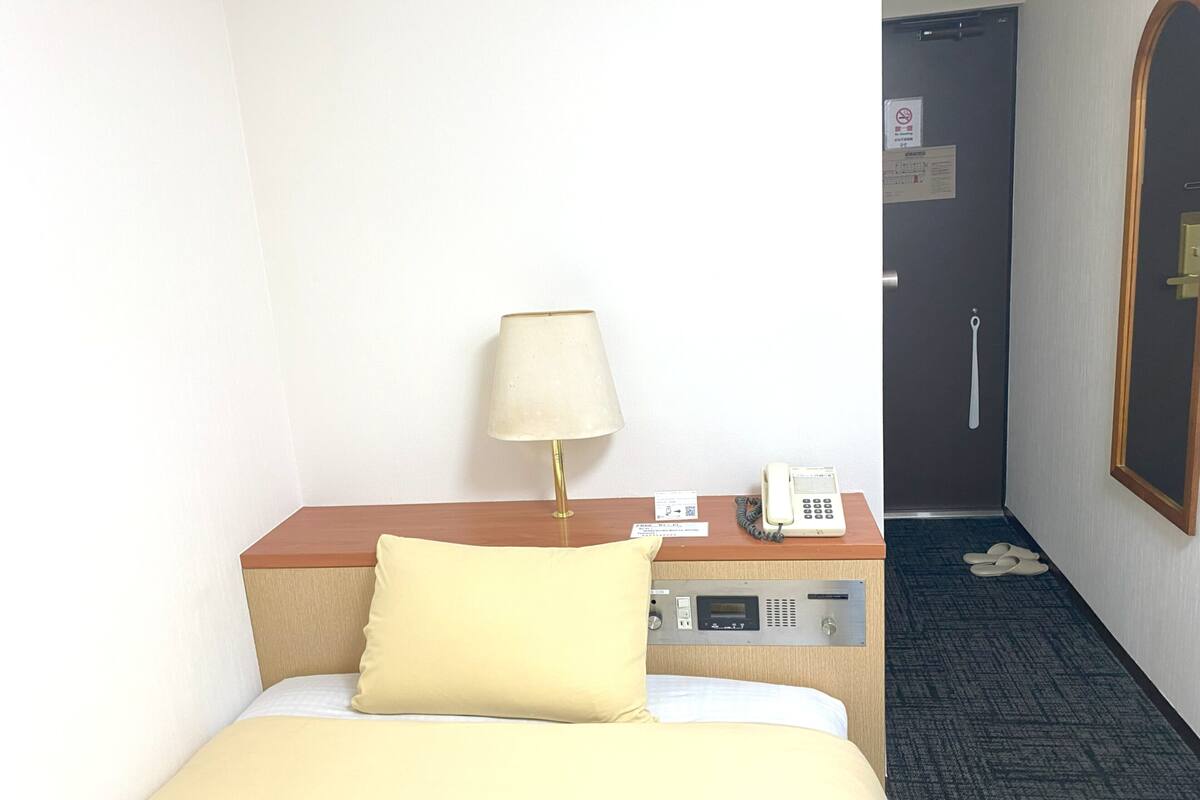 A hotel room with a bed, a bedside table with a lamp, a phone, and a door with a handle.